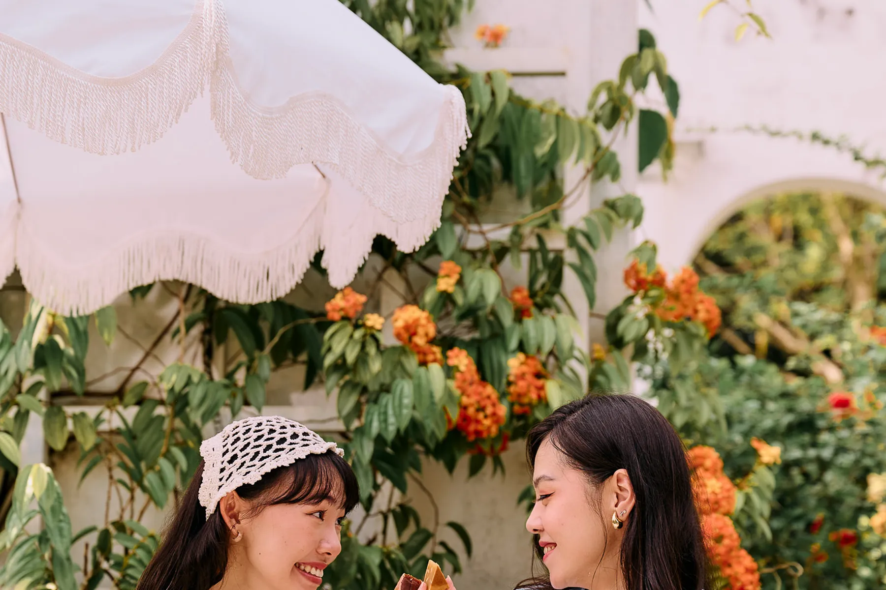 Two friends sharing mooncakes at a garden table beneath a fringed parasol
