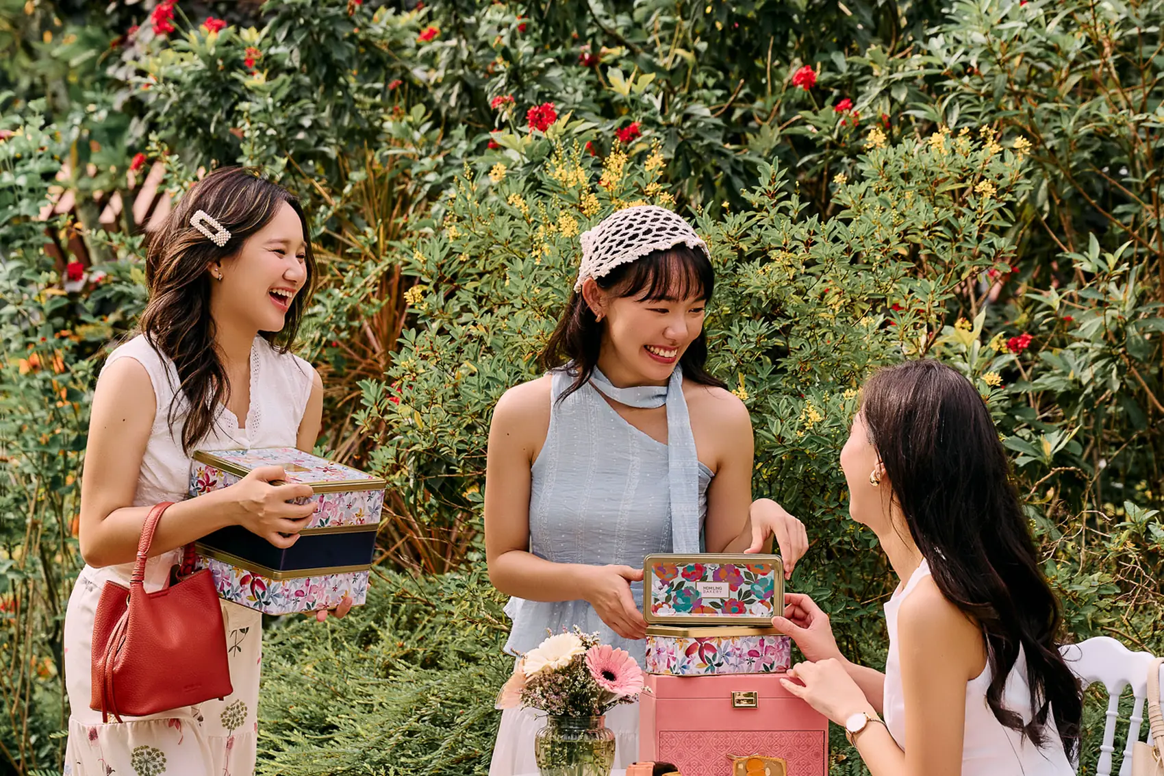 Three friends at a white garden table with Mdm Ling Bakery mooncake tins, in front of a flowering rose hedge